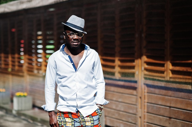 Elegante uomo afroamericano in camicia bianca e pantaloni colorati con cappello e occhiali in posa all'aperto Ragazzo nero modello alla moda