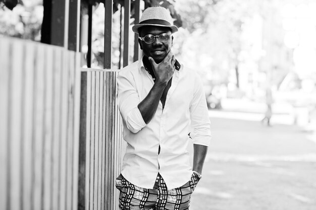 Elegante uomo afroamericano in camicia bianca e pantaloni colorati con cappello e occhiali in posa all'aperto Ragazzo nero modello alla moda