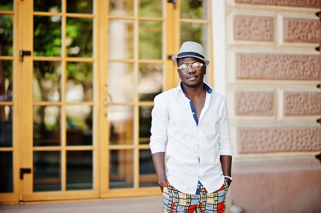 Elegante uomo afroamericano in camicia bianca e pantaloni colorati con cappello e occhiali in posa all'aperto Ragazzo nero modello alla moda