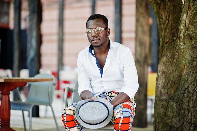 Elegante uomo afroamericano in camicia bianca e pantaloni colorati con cappello e occhiali in posa all'aperto Ragazzo nero modello alla moda