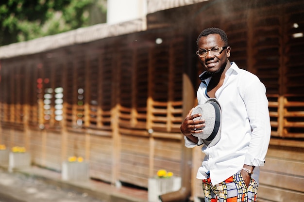 Elegante uomo afroamericano in camicia bianca e pantaloni colorati con cappello e occhiali in posa all'aperto Ragazzo nero modello alla moda