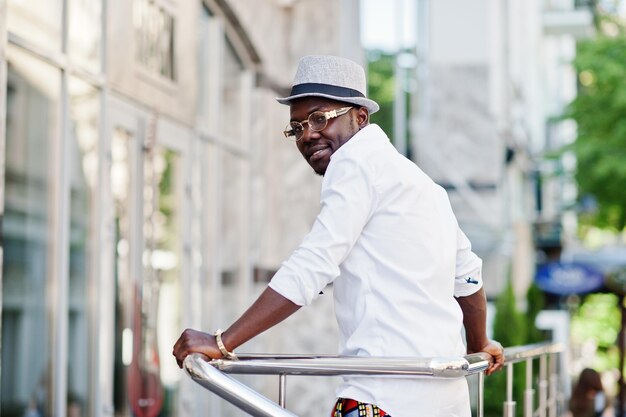 Elegante uomo afroamericano in camicia bianca e pantaloni colorati con cappello e occhiali in posa all'aperto Ragazzo nero modello alla moda