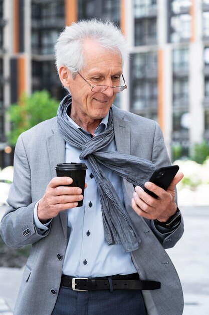 Elegante uomo anziano in città che usa lo smartphone mentre beve un caffè