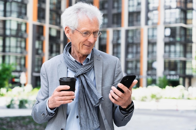 Elegante uomo anziano in città che usa lo smartphone mentre beve un caffè