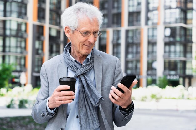 Elegante uomo anziano in città che usa lo smartphone mentre beve un caffè
