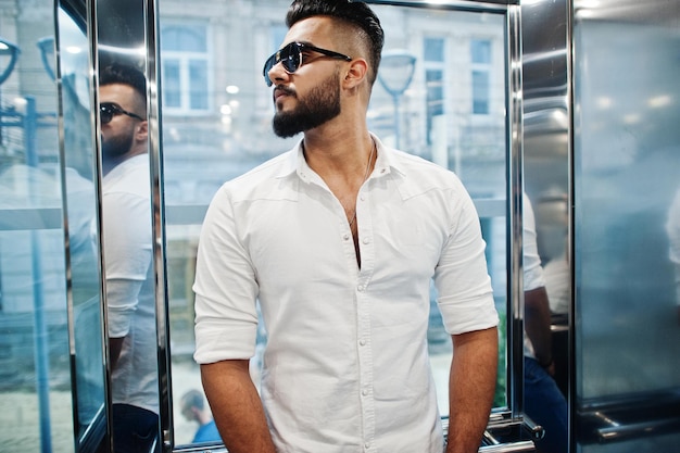 Elegante uomo arabo alto modello in camicia bianca e occhiali da sole posato all'interno dell'ascensore