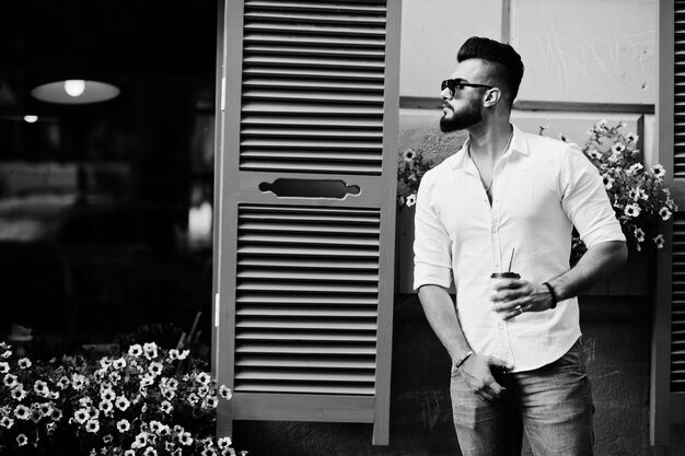 Elegante uomo arabo alto modello in camicia bianca, jeans e occhiali da sole posato in strada della città Barba attraente ragazzo arabo con una tazza di caffè contro le finestre viola
