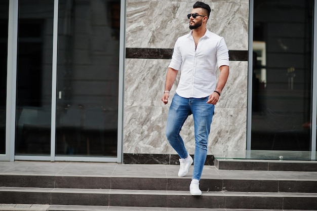 Elegante uomo arabo alto modello in camicia bianca jeans e occhiali da sole poste in strada della città Barba attraente ragazzo arabo