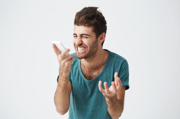 Elegante uomo barbuto con i capelli scuri che grida arrabbiato al telefono. Uomo arrabbiato e stressato, vestito con una maglietta blu che grida forte nella disperazione e nella rabbia, furioso per il rumore proveniente dal telefono