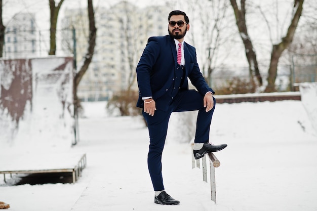 Elegante uomo d'affari con barba indiana in giacca e occhiali da sole in posa durante la giornata invernale all'aperto