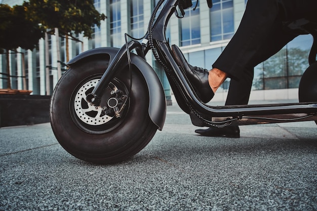 Elegante uomo d'affari in abito nero è seduto sul suo scooter elettrico vicino al suo ufficio.