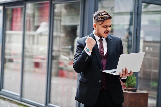 Elegante uomo d'affari indiano felice eccitato in abbigliamento formale con laptop sulle mani in piedi contro le finestre nel centro business Gesto del vincitore