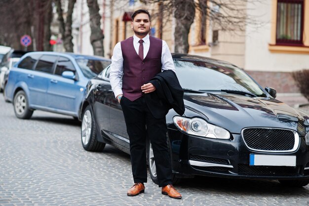 Elegante uomo d'affari indiano in abito formale gilet in piedi contro un'auto nera d'affari sulla strada della città