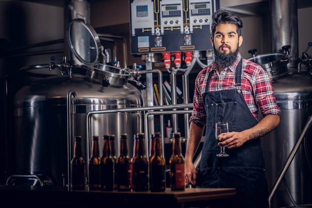 Elegante uomo indiano con la barba completa in una camicia in pile e grembiule tiene un bicchiere di birra, in piedi dietro il bancone in una fabbrica di birra.