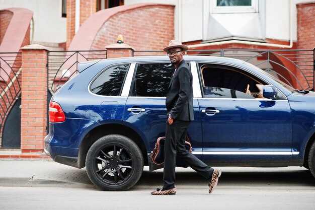 Elegante uomo nero con occhiali con cappello da indossare su tuta con borsetta contro auto di lusso Ricco uomo d'affari afroamericano