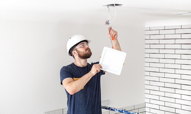 Elettricista Builder con barba lavoratore in un casco bianco al lavoro, installazione di lampade in altezza. Professionista in tuta con un trapano sul sito di riparazione.