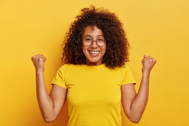 Emotiva donna dalla pelle scura fa il gesto evviva, alza i pugni, sorride piacevolmente, sorride divertito, indossa grandi occhiali rotondi e maglietta casual, ha i capelli ricci luminosi, isolato su sfondo giallo