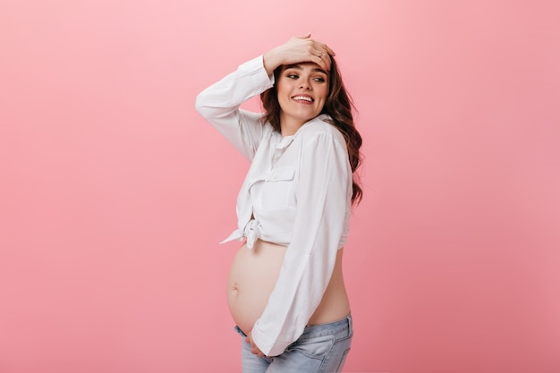 Emotiva donna incinta felice in jeans e camicia bianca ritagliata tocca i capelli e sorrisi su sfondo rosa isolato.