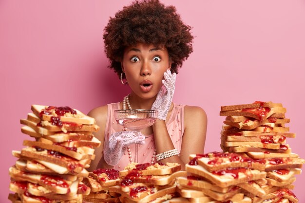 Emotiva donna stupefatta con capelli afro, fissa con occhi spalancati, tiene in mano un bicchiere di cocktail, vestita elegantemente, modelle contro un muro roseo, circondate da gustosi toast di pane, celebra qualcosa