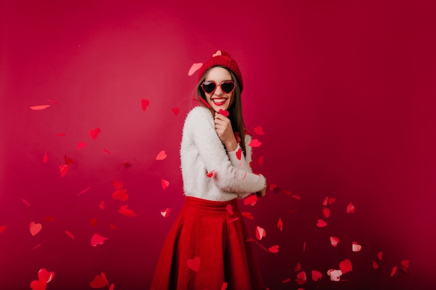 Emotiva giovane donna in cappello rosso e occhiali da sole in piedi sullo spazio bordeaux alla festa