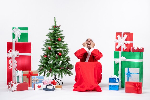 Emotivo eccitato interessato giovane uomo vestito da Babbo Natale con doni e albero di Natale decorato su sfondo bianco stock photo