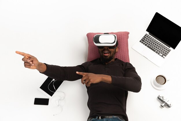 Emotivo uomo afro-americano utilizzando VR-auricolare circondato da gadget isolati su sfondo bianco studio, tecnologie. Gioco emotivo