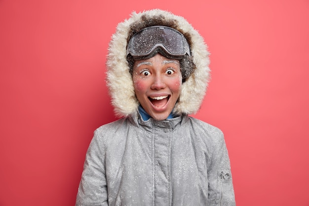 Emozionante ragazza invernale allegra guarda con un'espressione felicissima ha la faccia rossa coperta dalla brina ama fare snowboard durante il periodo invernale indossa una giacca calda.