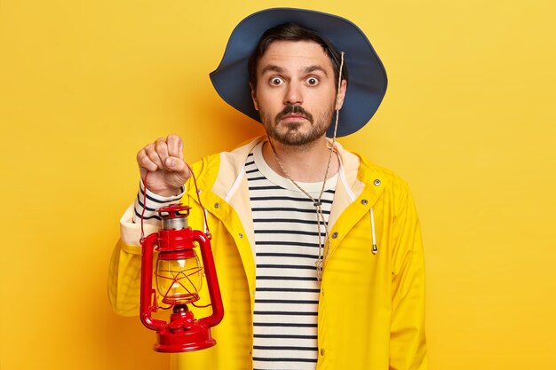 Emozionante ragazzo con la barba lunga indossa cappello, maglione a righe e impermeabile, tiene in mano una lampada a cherosene, trascorre il fine settimana nella natura, gode di una sosta notturna