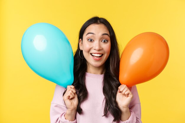 Emozioni delle persone, tempo libero lifestyle e concetto di bellezza. La ragazza asiatica felice allegra si congratula con la grande festa, tenendo in mano due palloncini e sorridendo ampiamente, godetevi la festa su sfondo giallo.