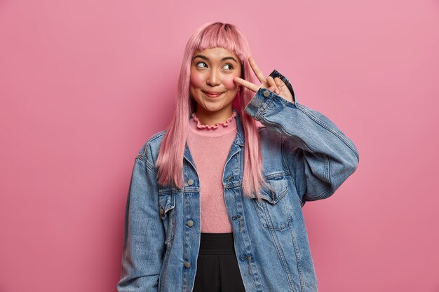 Emozioni positive e concetto di atteggiamento. La ragazza dai capelli rosa sognante sorridente dà il segno di vittoria di pace, mostra due dita