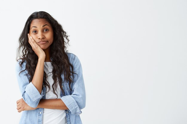 Emozioni, sentimenti, reazioni e atteggiamento umani. Attraente ragazza dalla pelle scura in camicia di jeans con lunghi capelli ondulati che tiene la mano sulla guancia in dubbio e sospetto, sentendosi scettica su qualcosa.