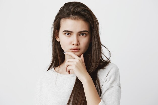 Emozioni, sentimenti, reazioni e atteggiamento umani. Bella donna in maglietta casual con lunghi capelli lisci scuri, tenendo la mano sul mento in dubbio e sospetto, sentendosi scettica su qualcosa