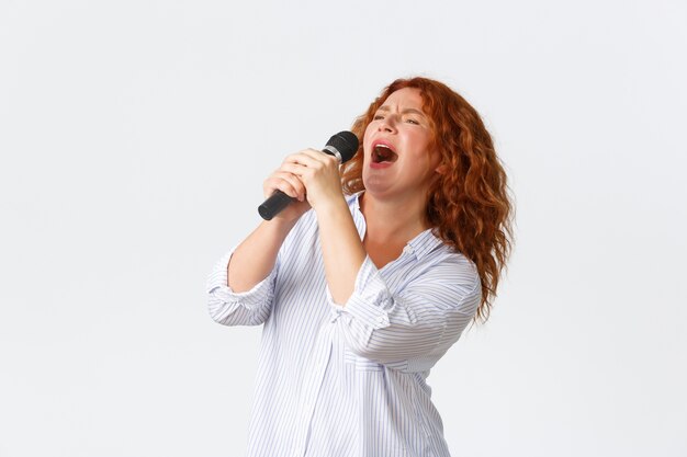 Emozioni, stile di vita e concetto di svago. Interprete femminile rossa appassionata e spensierata, donna di mezza età che canta una canzone nel microfono, cantante che gioca a karaoke, sfondo bianco