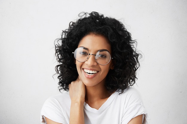 Emozioni umane positive. Ritratto di bella e affascinante studentessa con acconciatura afro, con sguardo timido, ridendo, indossando eleganti occhiali rotondi, toccando il collo con la mano