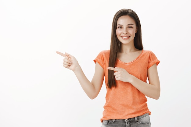 Entusiasta bella ragazza bruna che punta il dito a sinistra, mostrando fantastico promo, invitando a dare un'occhiata, dimostrare banner