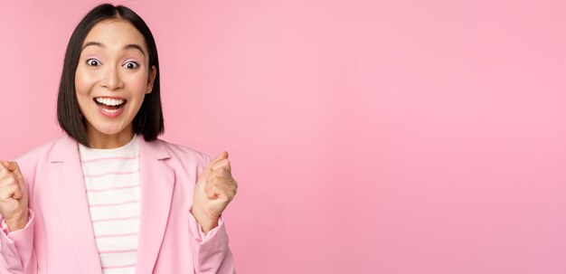 Entusiasta commessa asiatica donna aziendale dire di sì raggiungere l'obiettivo e celebrare trionfante guardando con gioia e sorridente in piedi su sfondo rosa