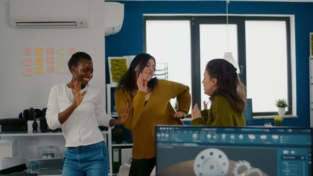 Entusiasta diverse donne ingegneri che ballano nell'ufficio aziendale celebrando il successo del progetto cantando