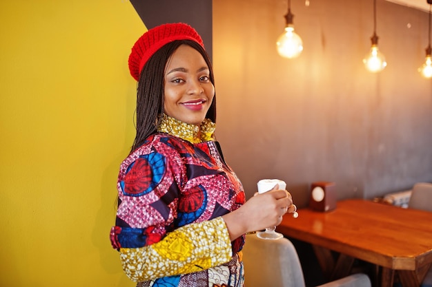 Entusiasta donna afroamericana in abito colorato alla moda con berretto rosso che si rilassa in un accogliente caffè con una tazza di latte caldo in mano