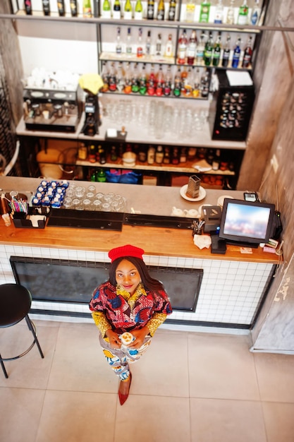 Entusiasta donna afroamericana in abito colorato alla moda con berretto rosso che si rilassa in un accogliente caffè in piedi vicino al bancone del bar