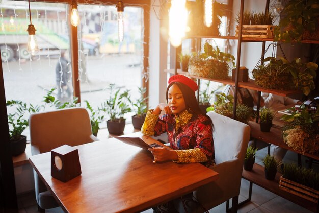 Entusiasta donna afroamericana in abito colorato alla moda con berretto rosso che si rilassa nell'accogliente menu di lettura del caffè