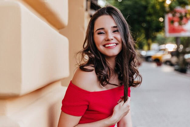 Entusiasta donna bianca che gioca con i suoi capelli ondulati mentre si trovava per strada. Modello femminile di buon umore in abito rosso in posa in città con un sorriso.