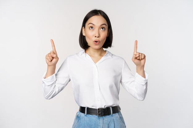 Entusiasta donna d'affari asiatica che punta guardando in alto con la faccia sorridente felice che mostra il logo dell'azienda o un banner in piedi su sfondo bianco