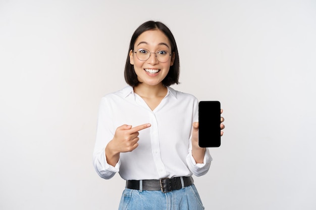 Entusiasta giovane donna asiatica che punta il dito sullo schermo dello smartphone che mostra la pubblicità su sfondo bianco del telefono cellulare