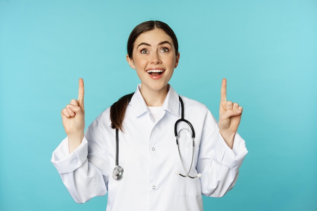 Entusiasta lavoratore medico, giovane donna medico in camice bianco, stetoscopio, mostrando pubblicità, puntando il dito verso l'alto, in piedi su sfondo turchese