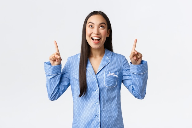 Entusiasta ragazza asiatica attraente in pigiama blu che punta le dita in alto e sorride stupita mentre guarda l'incredibile offerta di banner promozionali, mostra l'annuncio, reagisce al fantastico nuovo prodotto.