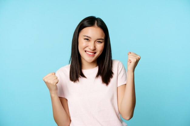 Entusiasta ragazza asiatica carina che vince, gioisce per le grandi notizie, danza della vittoria, sorride compiaciuta, in piedi su sfondo blu