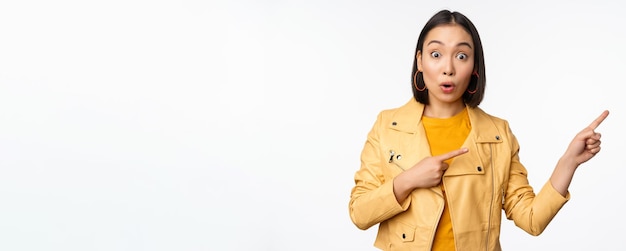 Entusiasta ragazza asiatica che punta il dito verso destra mostrando pubblicità e sorridente invitando al negozio in piedi su sfondo bianco