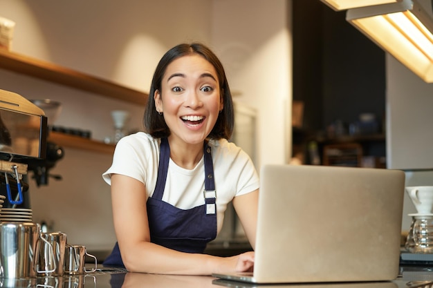 Entusiasta ragazza asiatica in un lavoratore barista uniforme da caffè con un computer portatile che sembra felice e sorpreso a ca