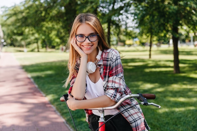 Entusiasta ragazza caucasica in occhiali carino seduto sulla bicicletta. Foto all'aperto di gioiosa modello femminile in posa con piacere nel parco verde.