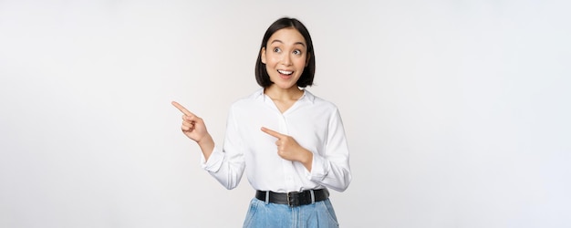 Entusiasta ragazza coreana che punta il dito a sinistra studentessa che punta e guarda a sinistra con un sorriso felice che mostra il logo dell'azienda o un banner sfondo bianco